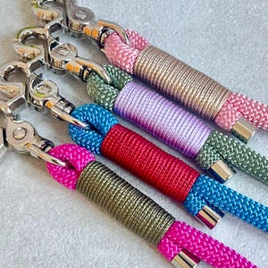 May include: Four keychains featuring silver-toned metal swivel lobster clasps attached to braided nylon cords. Each cord displays a two-tone color scheme, including combinations of pink and beige, olive green and lavender, blue and red, and hot pink and olive.