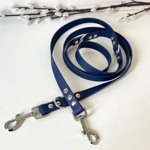 May include: A navy blue waterproof lead featuring a smooth, flexible strap with silver-toned metal hardware, including two swivel snap hooks and multiple D-rings secured by silver rivets, designed for versatile handling and secure attachment.