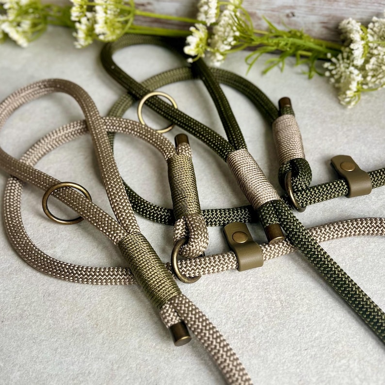 May include: Two braided rope leashes, one in a light beige and one in a dark olive green. Each leash features a circular metal ring, a matching metal end cap, and a secure metal rivet fastener connecting the rope segments in a durable design.