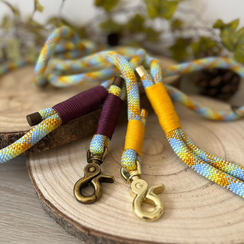 May include: Two braided rope leashes featuring a light blue and yellow diamond pattern. One leash has dark burgundy cord wrapping and an antique brass swivel snap hook, while the other has bright yellow cord wrapping and a polished gold-tone hook.