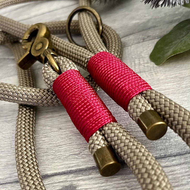 May include: A beige braided rope leash featuring two sections wrapped in vibrant pink cord. The leash includes antique brass hardware, specifically a swivel snap hook and a circular ring, with matching brass end caps on the rope segments.