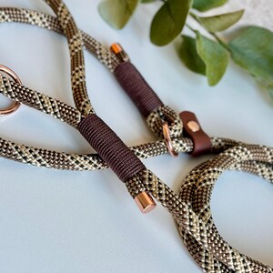May include: A dog leash made of braided rope in shades of brown, tan, and black. The leash features dark brown cord wrappings, rose gold metal rings, and rose gold metal end caps. A small brown leather tab with a rose gold rivet is attached.