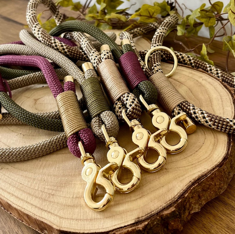 May include: A collection of braided rope leashes in olive green, plum, and tan patterns. Each leash features gold-toned metal swivel snap hooks and matching gold-toned cord wraps securing the rope ends. The ropes are textured and woven.