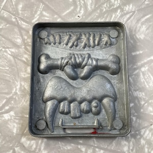 May include: A silver metal mold for making chocolate or candy. The mold features a bone, a set of teeth, and a stitched-up wound. The mold is marked with the letters "MD".