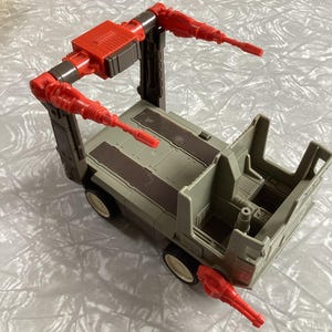 May include: A retro toy vehicle in grey and red. The toy has a truck bed, a cab, and a red articulated arm with nozzles. The wheels are off-white. The toy is likely from the 1980s.