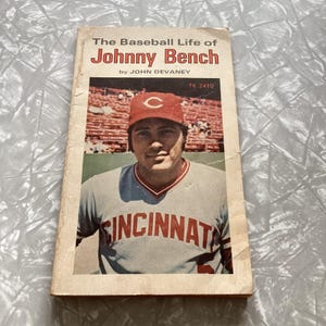 May include: A vintage book titled "The Baseball Life of Johnny Bench" by John Devaney. The cover shows a colour photograph of Johnny Bench in a Cincinnati Reds baseball uniform and cap. The book has a worn appearance.