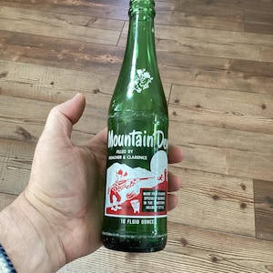 May include: A vintage green glass Mountain Dew bottle. The label features a cartoon illustration of a man with a rifle and two pigs. The bottle is labeled "10 fluid ounces".