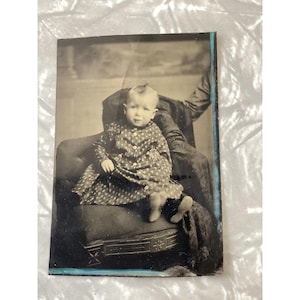 May include: A vintage black and white photograph featuring a young child seated on an upholstered chair. The child wears a patterned dress with light-colored buttons. A portion of an adult arm and clothing is visible on the right side of the frame.