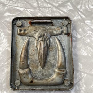 May include: A vintage metal mold for making chocolate candy. The mold features a detailed design of a mythical creature with horns and a beard. The mold is slightly tarnished and shows signs of age.