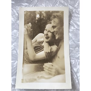 May include: Vintage black and white photograph of a laughing couple. The woman is wearing a striped top and the man is shirtless. They are embracing and appear to be enjoying a moment of joy.