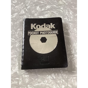 May include: A black Kodak Pocket Photoguide book with silver text and a silver camera aperture graphic on the front cover. The title Kodak Pocket Photoguide is printed in bold silver lettering above a circular camera shutter design.