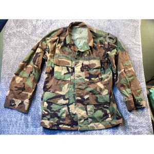 May include: Camouflage military jacket with multiple pockets and the name "Bussell" above the left pocket. The jacket also has "U.S. ARMY" above the right pocket. The jacket is in shades of green, brown, and black.