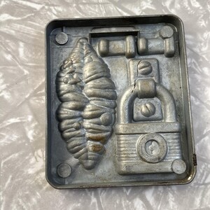May include: A vintage metal mold for making chocolate candies. The mold features three designs: a leaf, a padlock, and a door handle.