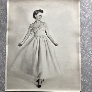 May include: A black and white photo of a woman wearing a white dress with a bow at the neckline. She is standing in a studio setting with a plain background.