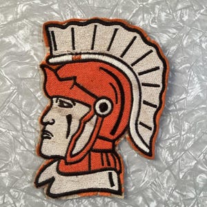 May include: Orange and white embroidered patch of a Roman soldier's head wearing a helmet with a plume.