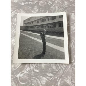Peut inclure: Une photographie en noir et blanc montrant une personne portant un casque et un uniforme de style militaire avec un sac à dos, debout sur un terrain herbeux devant un bâtiment à plusieurs étages avec des rangées de fenêtres et une structure.