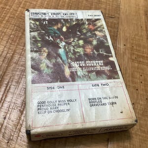 May include: A vintage cassette tape case with a photo of a band on the front. The case is labeled "Bayou Country" and features the band "Reedenne Clearwater Revival". The back of the case lists the songs on each side: "Good Golly Miss Molly", "Penthouse Pauper", "Proud Mary", "Keep On Chooglin'" and "Born On The Bayou", "Bootleg", "Graveyard Train".