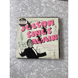 May include: Vintage vinyl record album cover for "Jolson Sings Again" by Decca Records. The cover features a smiling man in a tuxedo, with the title in pink letters. The album is a 45 RPM record.