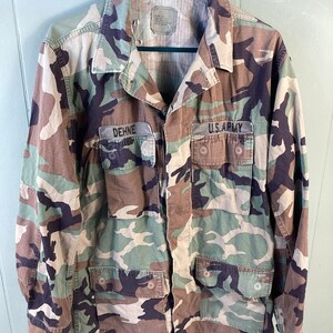 May include: Camouflage military jacket with the name "DEHNE" and "U.S. ARMY" patches. The jacket features a button-up front, multiple pockets, and a collar. The pattern includes shades of green, brown, and black.