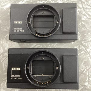 May include: Two black camera body parts with a silver lens mount and a silver label that reads "ZEISS West Germany 45 60 70 80".