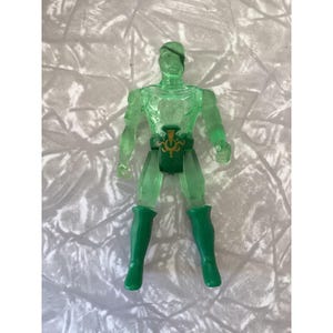 May include: A translucent green action figure with a muscular build. The figure has green boots and a green belt with a gold emblem. The figure is standing upright, with arms at its sides.