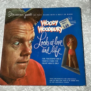 May include: A blue vinyl record cover with a photo of a man and a woman. The title of the record is "Woody Woodbury Looks at Love and Life". The text on the cover says "Fun, Foolosophy and a Frantic Piano for Frisky Adults Only". The record is from the Bahama Hotel in Fort Lauderdale, Florida.