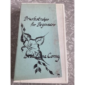 May include: A white VHS cassette tape with a light blue paper label. The label features black text that reads Brushstrokes for Beginners with Edna Corney and a black line drawing of a flower with leaves extending down the center of the cover.