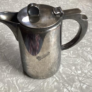 May include: A vintage silver-coloured metal coffee pot with a lid and handle. The pot features a pouring spout and the word "Butlin's" is inscribed on the side. It rests on a white, textured surface.