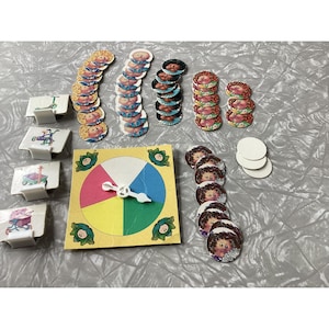 May include: A vintage board game featuring a colorful spinner, game pieces with character illustrations, and small square game cards with vehicle graphics. The game pieces are arranged on a light gray surface.