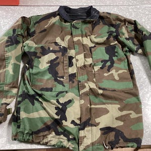 May include: Camouflage jacket in green, brown, and black. It has a black collar, two front pockets, and a drawstring at the hem. The long-sleeved jacket is made of a durable fabric. The pattern is a classic military design.