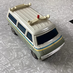 May include: A vintage toy van with a white roof and gray body. The van has a red light on top and a white roof rack. The van has yellow and blue stripes on the side.