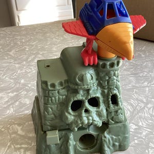 May include: A vintage toy playset featuring a green castle with a skull design and a blue and red bird-like vehicle. The toy is made of plastic and appears to be from a children's action figure line.