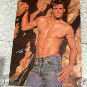 May include: A poster featuring a man with a muscular physique, wearing blue jeans. The background has a textured, brown rock formation. The name "Tom Wright" is signed in the upper left corner.