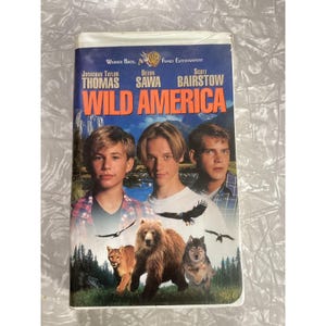 May include: A VHS tape cover for the film Wild America featuring the actors Jonathan Taylor Thomas, Devon Sawa, and Scott Bairstow. The artwork displays a mountain landscape with a brown bear, a wolf, and a mountain lion in the foreground.