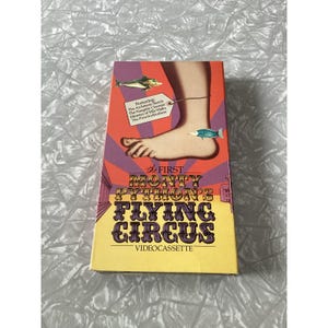 May include: A rectangular videocassette box featuring a graphic of a human foot with a tag attached, surrounded by small fish illustrations. The text reads: The First Monty Python's Flying Circus, Featuring: The Architects Sketch, The Naughty Chemist, Ministry of Silly Walks, The Piranha Brothers.