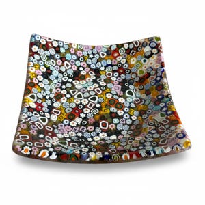 Op de afbeelding: Een vierkante glazen schaal met een dicht millefiori-patroon bestaande uit talloze kleine, kleurrijke bloemvormige en geometrische figuren in rood, geel, blauw, groen, wit en zwart, wat zorgt voor een levendige, mozaïekachtige textuur.