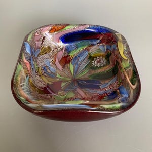 Peut inclure: Un bol décoratif en verre de forme carrée aux coins arrondis. Le bol présente un design multicolore vibrant avec des nuances de bleu, de rose, de vert et d'or. La base du bol est de couleur rouge foncé.