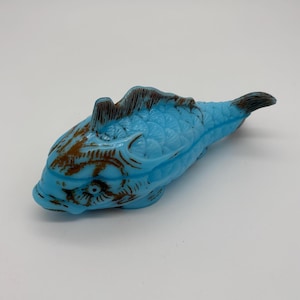 Pode incluir: Uma estatueta de peixe azul claro com detalhes em castanho. O peixe tem escamas e barbatanas detalhadas, com uma superfície texturizada. É um objeto decorativo.