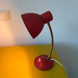 Op de afbeelding: Een rode bureaulamp met een koepelvormige kap, een flexibele metalen zwanenhalsarm en een ronde rode voet. De lamp is voorzien van een kleine witte drukknopschakelaar op de voet en een wit netsnoer dat uit de zijkant van de voet komt.