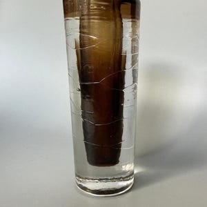 Può includere: Un vaso cilindrico in vetro caratterizzato da uno strato esterno trasparente con delicate e sottili linee bianche orizzontali e un nucleo interno color marrone scuro solido che si restringe leggermente verso il basso, creando un effetto visivo.