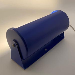 Könnte beinhalten: Eine blaue, zylindrische Wandleuchte mit rechteckigem Sockel. Die Lampe hat ein abgerundetes Ende und einen kleinen blauen Knopf oben. Ein weißes Kabel erstreckt sich von der Rückseite. Die Lampe ist für die Innenbeleuchtung konzipiert.