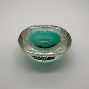 Könnte beinhalten: Eine klare Glasschale mit türkisem Innenraum. Die Schale hat eine runde Form und weist kleine, blasenartige Einschlüsse im Glas auf. Der Innenraum hat eine glatte, glänzende Oberfläche. Durchmesser ca. 10 cm.