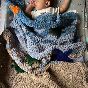 May include: A baby boy sleeping in a blue, brown, and green crocheted blanket with a star and a turtle design. The blanket is made of chunky yarn and has a soft, fluffy texture. The baby is wearing a white shirt with the word "WILD" printed on it.