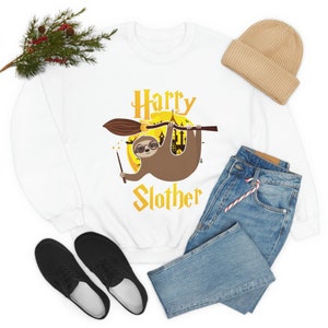 May include: White sweatshirt with a cartoon sloth wearing glasses and holding a wand, riding a broom. The text "Harry Slother" is in gold lettering. A tan beanie, blue jeans, and black shoes are also in the image.