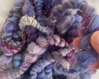 May include: A bundle of purple, blue, and white yarn with silver thread woven throughout. The yarn is soft and fluffy, and it is perfect for knitting or crocheting.
