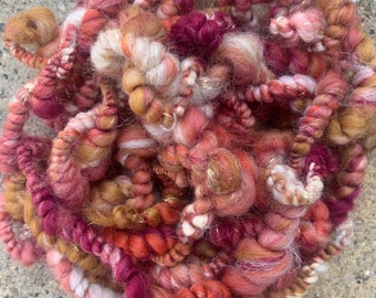 May include: A tangled mass of hand-spun yarn in shades of pink, rose, peach, and gold. The yarn has a thick, textured appearance with varying thicknesses and a subtle metallic sheen. Ideal for fiber art projects.