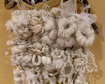 May include: A textured, off-white fiber art wall hanging. The piece is crafted with various yarns, ribbons, and embellishments, including pearls, lace, and a sheer bow. It is suspended from a natural driftwood branch with a hanging cord.