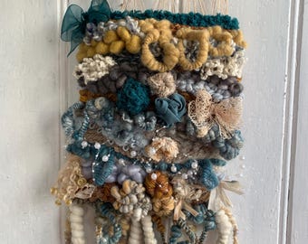 Könnte beinhalten: Ein gewebter Wandbehang mit einem Treibholzaufhänger. Der Behang zeigt eine Vielzahl von Texturen und Farben, darunter Blau, Türkis, Weiß, Beige und Gelb.