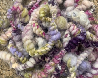 May include: A pile of colorful yarn in shades of purple, pink, green, and white. The yarn is twisted and knotted, creating a textured and fluffy appearance.