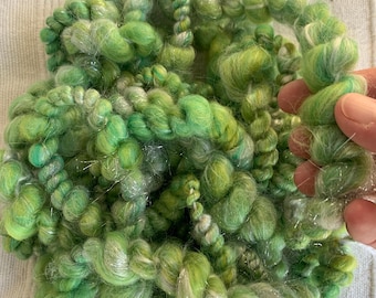 May include: A skein of green and white yarn with silver thread woven throughout. The yarn is fluffy and textured, with a hand-spun look.